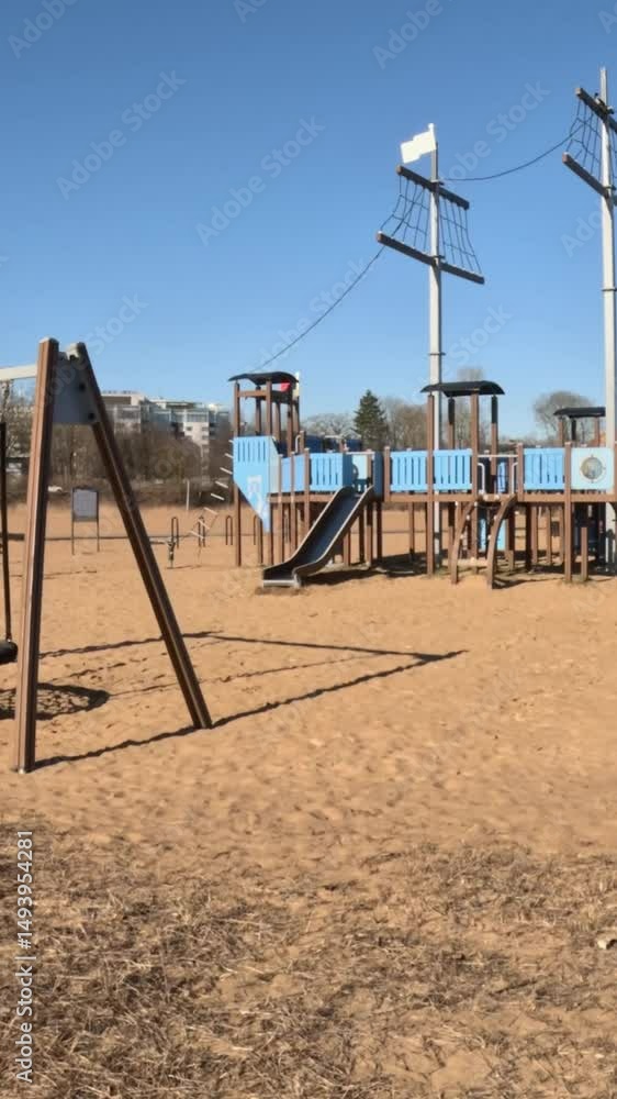 Wide shot of an empty playground featuring a large pirate ship playset and swings on sandy ground under a clear blue sky. Wide shot of an empty playground featuring a large pirate ship playset and swings on sandy ground under a clear blue sky.