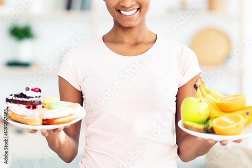 Tableau sur toile Lovely black lady holding plates with fruits and sweets, looking at unhealthy pastries, tempted to eat them at home