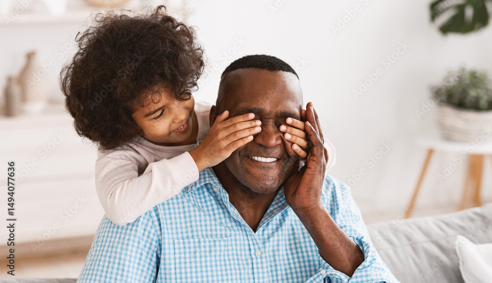 Generations bonding concept. Adorable African American girl closing her granddad's eyes at home, playing GUESS WHO game Generations bonding concept. Adorable African American girl closing her granddad's eyes at home, playing GUESS WHO game