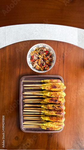 Indonesian satay lilit is served on skewers with traditional sambal matah. It is a delicious Balinese dish presented on a wooden table in a top-down view.