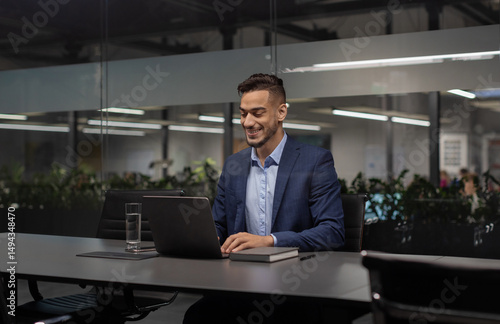 Positive young arabic manager working on laptop at modern hi-tech office, sitting at table, typing on computer keyboard, sending emails, communicating with business partners, copy space