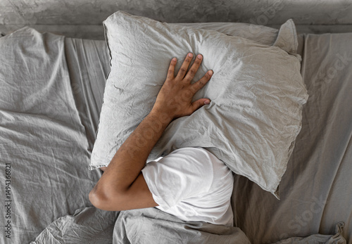 Tablou pe pânză Closeup of irritated young black man lying in bed and covering head and ears with pillow, hearing and suffering from too loud sound