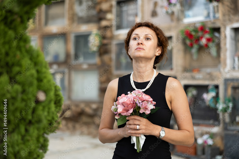 Fototapeta premium Tragic moment of visiting cemetery worshiping memory of deceased relative. Thoughtful and sad woman in black clothes with bouquet of hydrangeas mourns dead friend, stands near tombstone in solitude