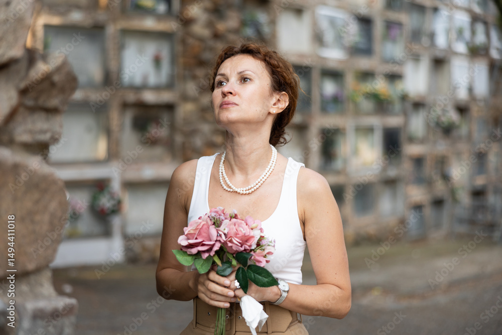 Fototapeta premium Elegant grieving woman in forties, wearing white tank top and pearls, standing with bouquet in hands near stone columbarium wall in cemetery, gazing sorrowfully and contemplating loss