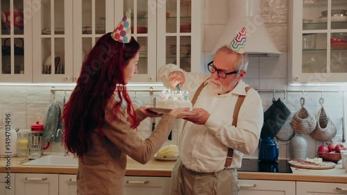 Grandpa holds a lighter with which he lights the candles on the cake which his daughter is holding standing opposite, then they carry it to grandma who blows them out