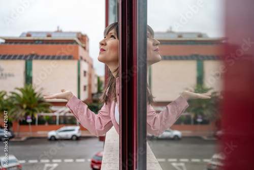 Woman leaning on balcony railing reflecting in window and looking up with open arms