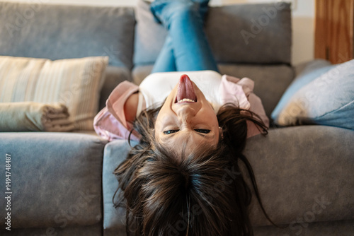 Young woman lying upside down on sofa sticking out tongue