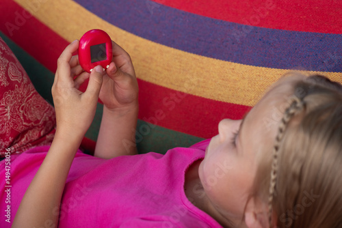 Little girl playing Tamagotchi toy while lying on a striped hammock. Popular game, caring for a virtual pet in the 90s.