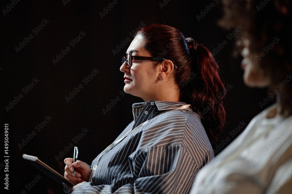 © Seventyfour - Side view of concentrated Middle Eastern female finance professional writing key points of presentation in notebook, while attending executive leadership event in auditorium with contrasting lighting