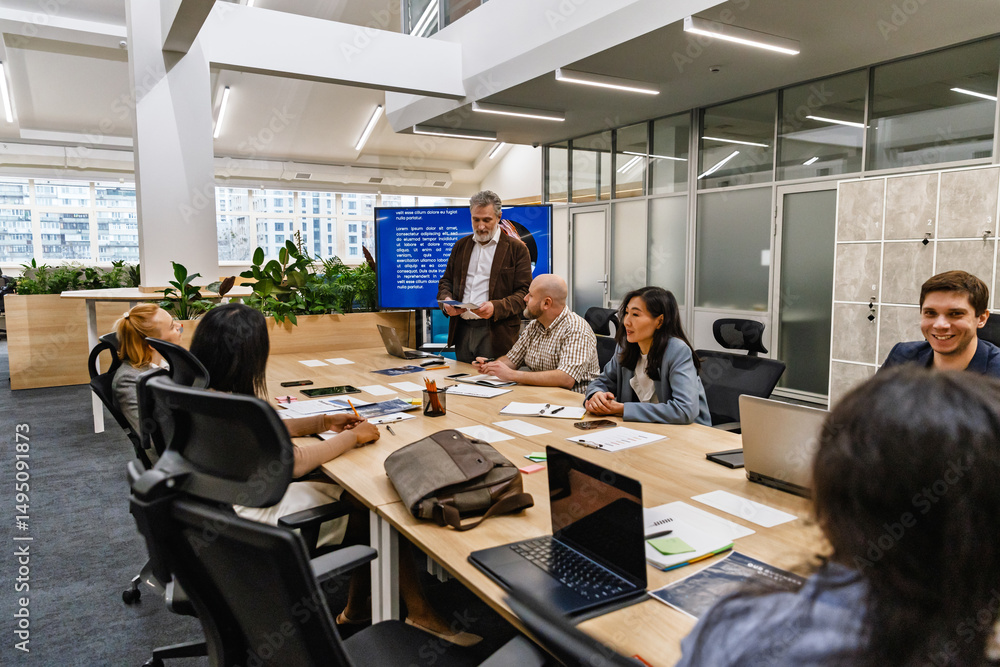 A male worker stands and looks at the papers he is holding while a group of six workers sit at a table next to him, in a meeting A male worker stands and looks at the papers he is holding while a group of six workers sit at a table next to him, in a meeting