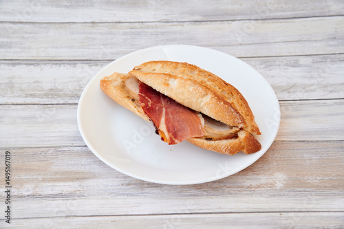 A ham sandwich is on a white plate. The sandwich is cut in half. The plate is on a wooden table