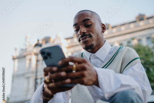 Obraz na plátně focused Black man in a preppy outfit sits outdoors, scrolling on his smartphone