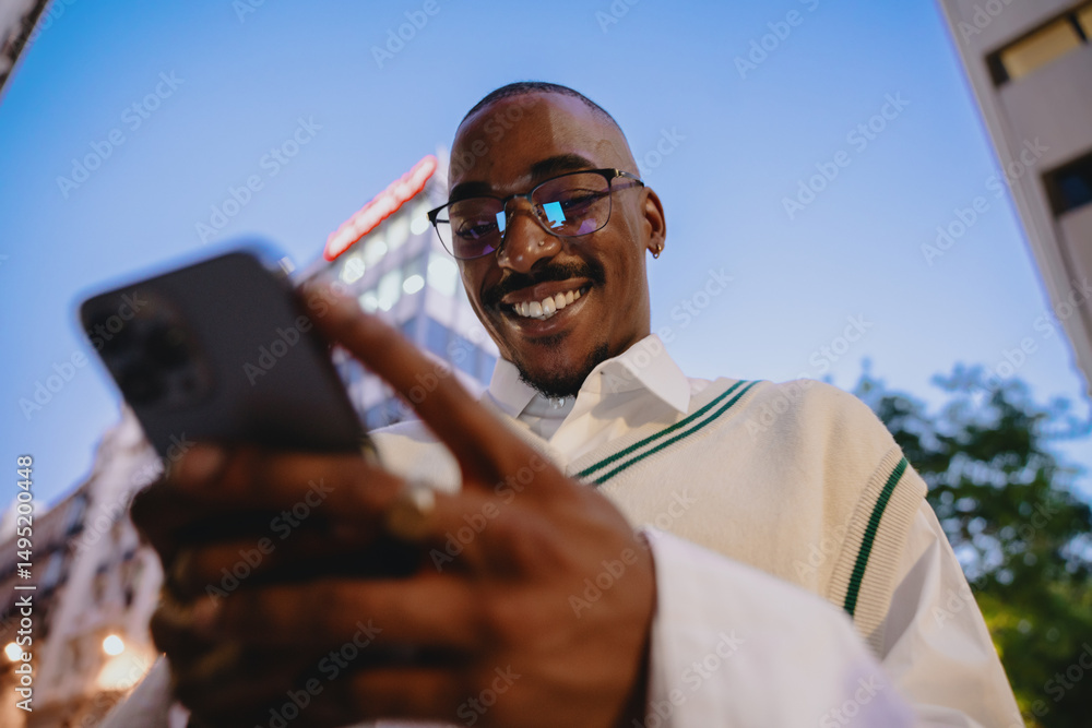 © BullRun - close-up of a young Black happy man in glasses looking at smartphone app with bright smile, reflecting city lights at night. His confident expression and trendy outfit add to modern urban aesthetic © BullRun - close-up of a young Black happy man in glasses looking at smartphone app with bright smile, reflecting city lights at night. His confident expression and trendy outfit add to modern urban aesthetic