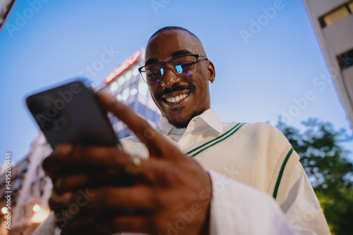Quadro su tela close-up of a young Black happy man in glasses looking at smartphone app with bright smile, reflecting city lights at night