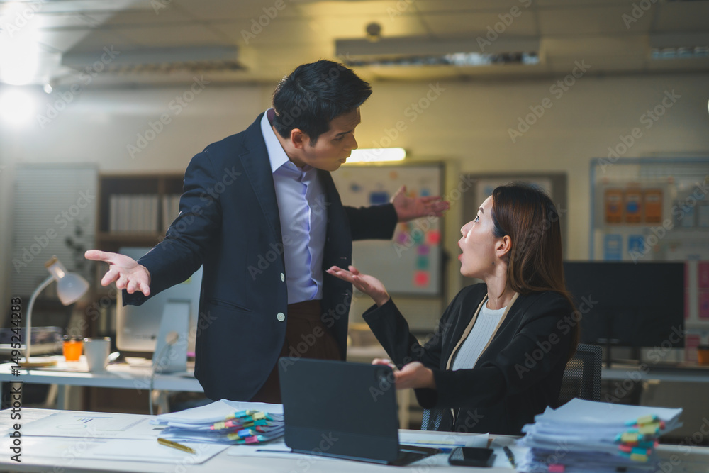 Fototapeta premium Two Asian businesspeople arguing about work while seated at a desk in an office, facing the stress and pressure of a looming deadline at night