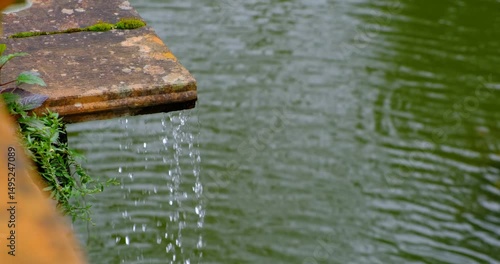 Water gently trickles from a weathered, moss-covered stone ledge into a serene green pond below. The scene is framed by delicate foliage, capturing quiet charm timeworn surfaces and nature’s calm flow