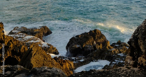 Waves crash with force against the jagged coastal rocks of Cascais, Portugal, under the warm glow of golden hour. Sea spray fills the air as the Atlantic puts on a dramatic, untamed display of power