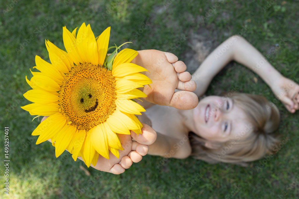 cheerful child lies on the grass, raising bare feet up, holding a sunflower with a carved smile. Focus on a large flower. Summer mood, floral fantasies, atmosphere of positivity and fun. cheerful child lies on the grass, raising bare feet up, holding a sunflower with a carved smile. Focus on a large flower. Summer mood, floral fantasies, atmosphere of positivity and fun.