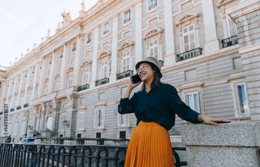 Fototapeta premium Smiling young Asian woman in hat and glasses talking on smartphone while traveling in Madrid, enjoying summer vacation, roaming service, and cultural cityscape near historic architecture