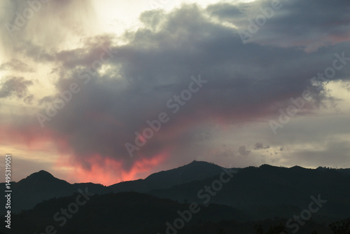 Dark mountain silhouettes beneath dramatic clouds glowing with fiery red and pink hues during a dramatic, moody sunset.