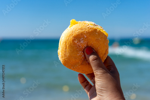 A woman’s hand holding a traditional Portuguese Bola de Berlim against the vibrant blue sea, capturing a perfect summer beach moment.