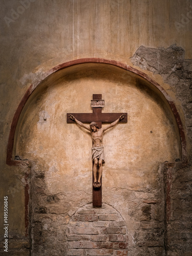 A wooden crucifix depicting Jesus Christ is mounted on an arched, weathered stone wall inside a historic church in Grado, Italy. The worn textures reflect time and reverence.