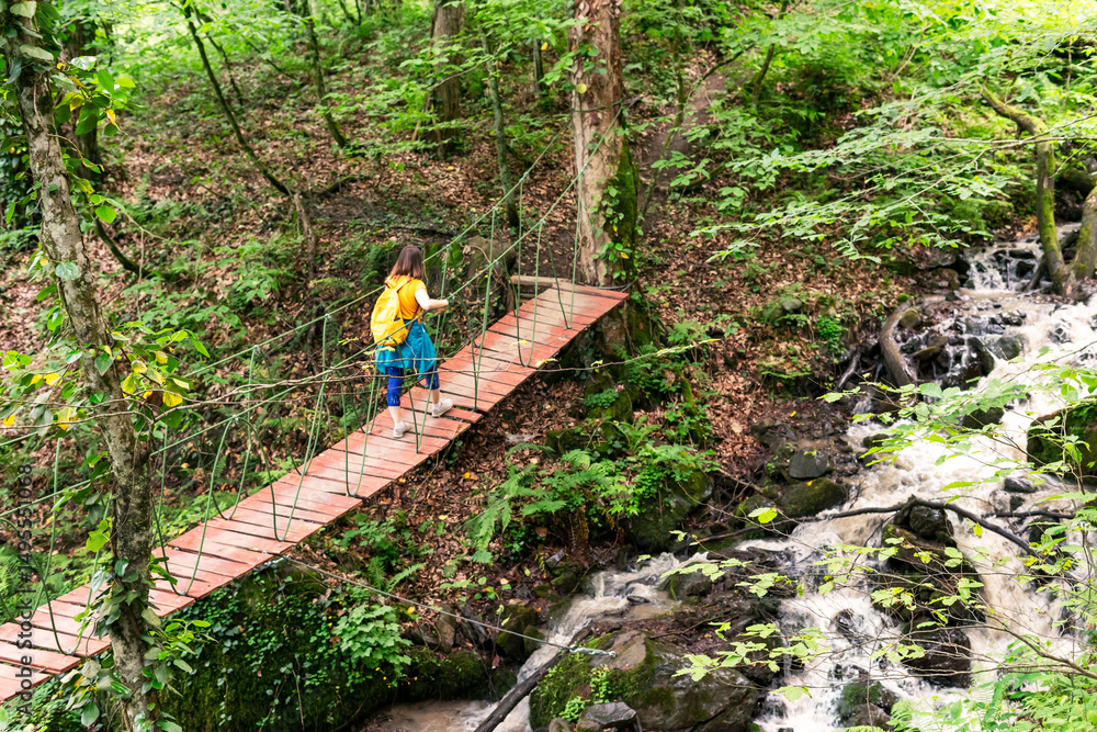 Naklejka premium Female hiker with yellow backpack walking on a wooden rope bridge over a stream in a green forest, adventure and travel concept