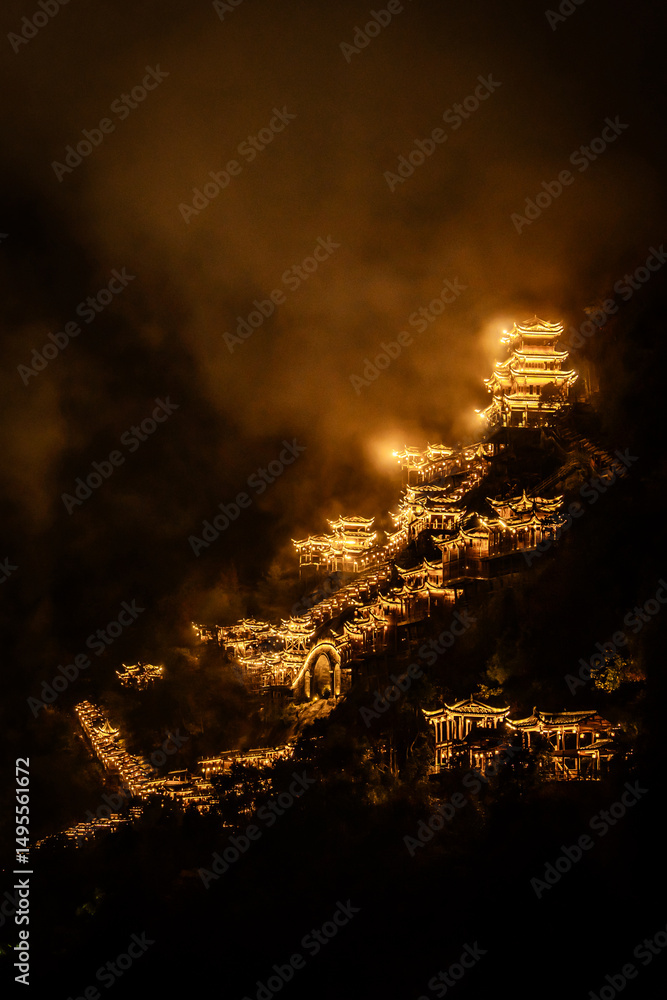 Fototapeta premium Aerial night view of Wangxian Valley Town, showcasing glowing traditional architecture nestled in the mountains, surrounded by nature and warm ambient lights.