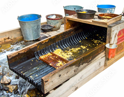 A traditional gold mining setup featuring a sluice box with water flow, surrounded by buckets, in a natural outdoor setting