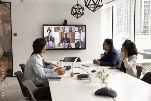 Group of multiethnic professionals sit at conference table, engaged in virtual meeting, looking at screen displaying diverse videoconference participants. Digital collaboration, remote communication