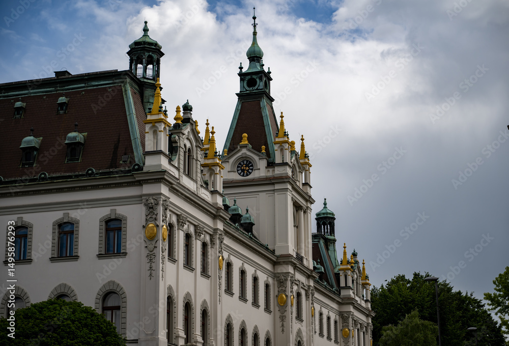 Fototapeta premium Ornate historical building with intricate architecture and gold accents, set against a cloudy afternoon sky with lush greenery in the foreground.