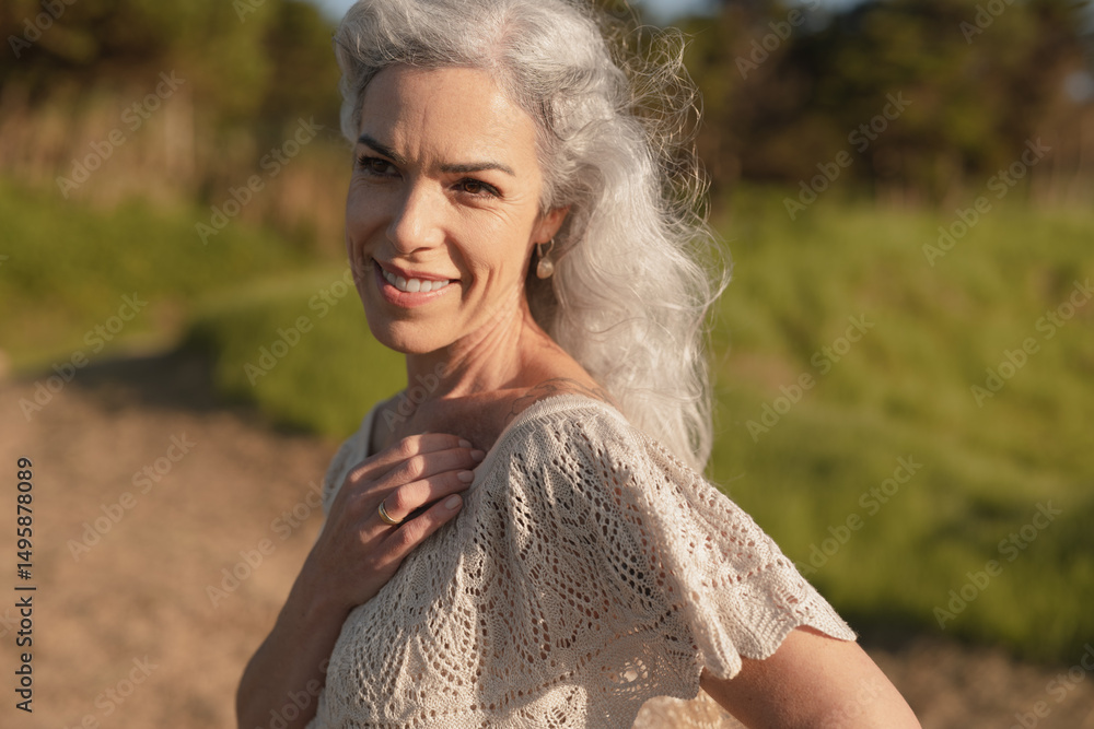 Naklejka premium A joyful older woman with silver hair smiles warmly outdoors, basking in sunlight. Her relaxed expression and gentle pose capture a sense of tranquility and contentment in a natural setting.