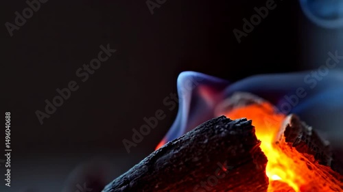 Close-up of burning wood with bright orange flames and flowing blue smoke against a dark background for abstract concepts or warm indoor visuals