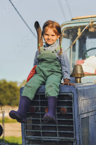 Vertical shot of farmer kid girl with two braids sitting on a tractor and smilling on a farm in summer. Happy childhood in the countryside. Country life