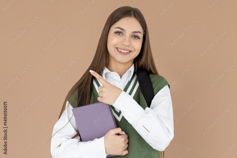 Fototapeta premium Hey look. Smiling pretty female model pointing fingers right at empty space and inviting to check it out, showing advertisement on brown background. Girl with notepads in hands.