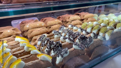 Assorted traditional Sicilian cannoli pastries filled with ricotta cream and decorated with chocolate chips, pistachios, and candied orange, displayed in a bakery glass case