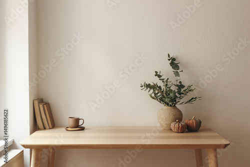 Autumn interior still life. Vase with eucalyptus, olive tree branches, pumpkins. Cup of tea, coffee, books. Wooden table at window. Home office. Minimal Scandi interior. Empty white wall background.