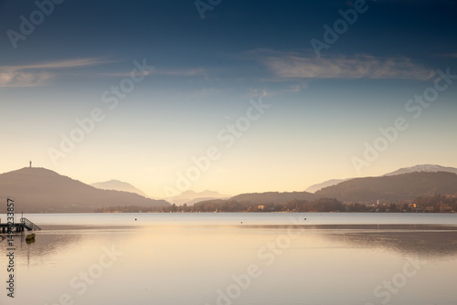 Wallpaper Mural Golden dusk settles over Lake Worthersee near Klagenfurt; Austria. calm mirror-like water reflects distant wooded hills and pale Alpine silhouettes beneath a deepening blue sky. Torontodigital.ca