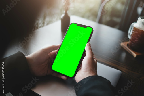 User holding smartphone with green screen at dark wooden desk with iced coffee in background, showcasing mobile app concept or user interface design.