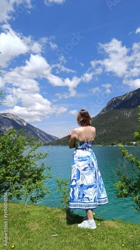 Woman in blue dress by the serene lake with mountains under a partly cloudy sky during summer afternoon