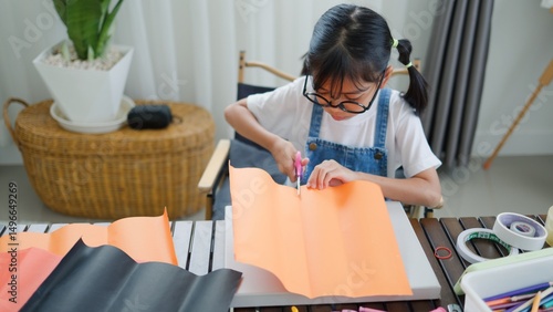 Fotografi Asian girl focused on making a DIY craft project at home using a compass and colorful paper, promoting creativity, fine motor skills, and fun learning in a homeschool setting