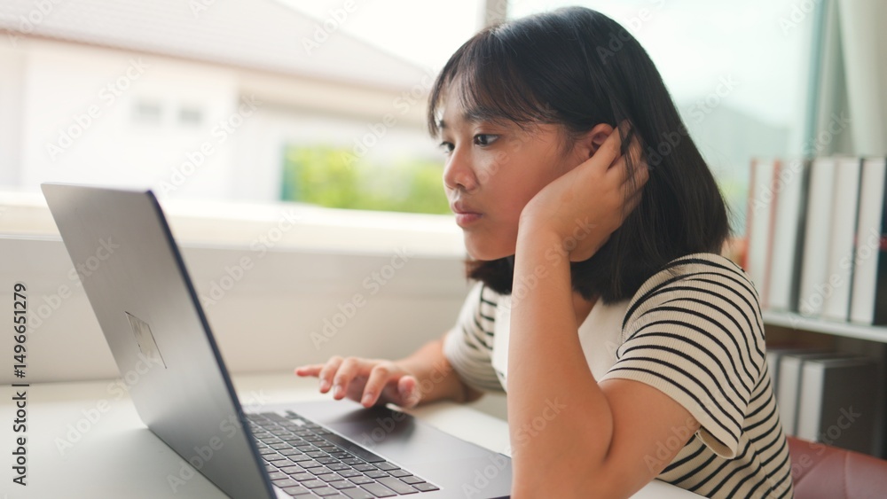 Asian teenage girl attentively watching an online class on a laptop at home, representing focused digital learning, homeschool, and remote education in a calm study space. Asian teenage girl attentively watching an online class on a laptop at home, representing focused digital learning, homeschool, and remote education in a calm study space.