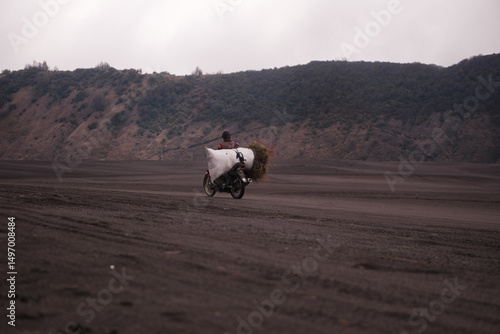 a person on a motorcycle through a volcanic environment, with a bundle of grass on the back. Java, Indonesia - 19th March 2025