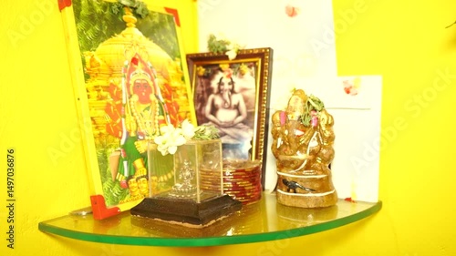 A vibrant home shrine with Hindu deities including Lord Ganesha, adorned with flowers. Colorful portraits and statues rest on a curved glass shelf against a bright yellow wall.