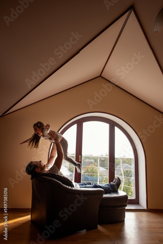 Father lifts his daughter joyfully in a cozy attic living room with a large arched window, capturing a warm and playful family moment filled with love and connection