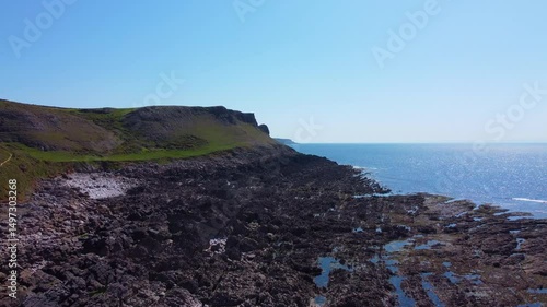 Slow Rising Drone Aerial View of Steep Cliff Tops with Bay at Low Tide and Sun Reflecting on Sea Water Creating Wide Picturesque View.