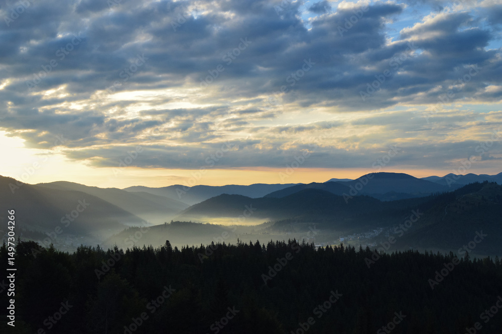 Sun rising over the Carpathian mountains, panoramic view. Silhouettes of mountain peaks. Morning fog. Dense forest in the foreground. Sun rising over the Carpathian mountains, panoramic view. Silhouettes of mountain peaks. Morning fog. Dense forest in the foreground.