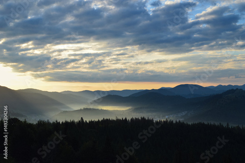 Sun rising over the Carpathian mountains, panoramic view. Silhouettes of mountain peaks. Morning fog. Dense forest in the foreground.