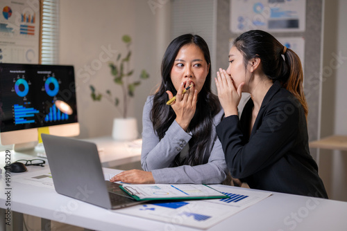 Two women are sitting at a desk with a laptop and papers in front of them