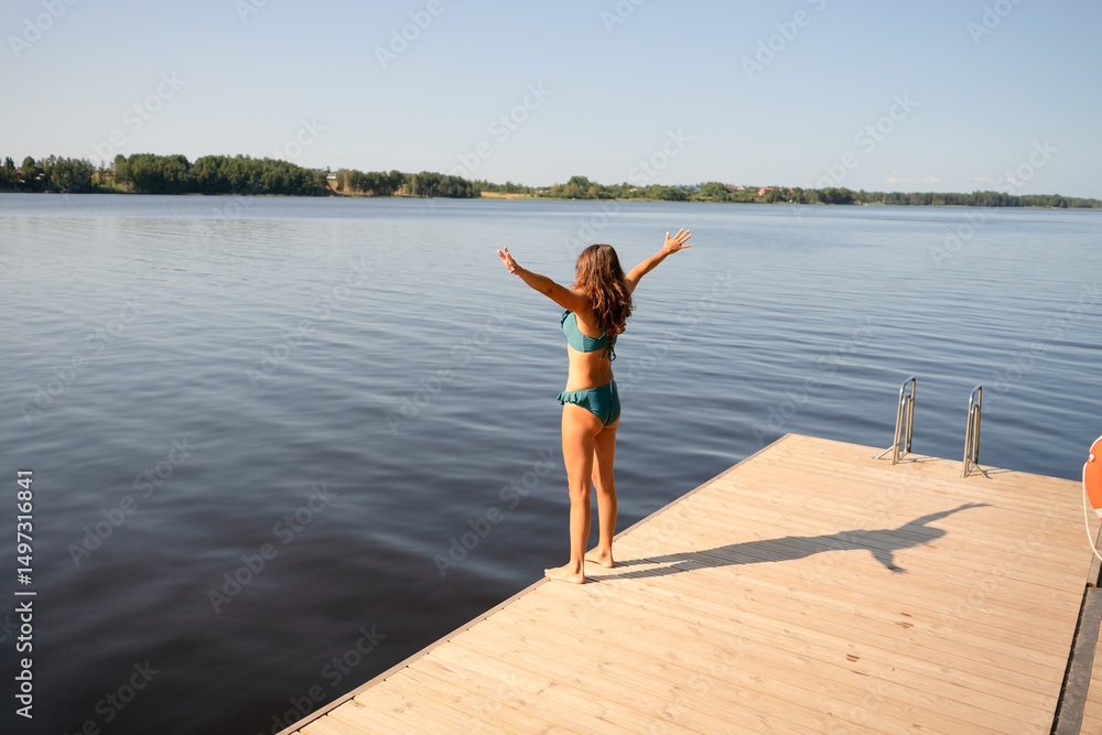 Obraz premium Woman in swimsuit standing on a wooden pier by the lake with arms wide open before swimming. Sunny weather, active lifestyle, and outdoor relaxation.